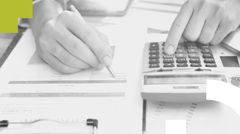 Foto en blanco y negro de una persona rellenando un documento con la ayuda de una calculadora.